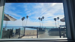 Vista a la playa desde una ventana, con palmeras, un muelle y sombrillas de playa.