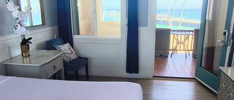Ett hotellrum med en stor säng, ett skrivbord, en stol, en takfläkt och utsikt över stranden.