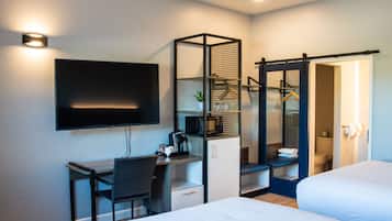 A modern hotel room with a flat-screen TV mounted on the wall, a desk with a chair, a bed, a wardrobe with a mirror, and a small kitchenette area.