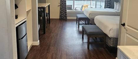 A compact hotel room with a bed, a small table, a bench, and a view of the outside.