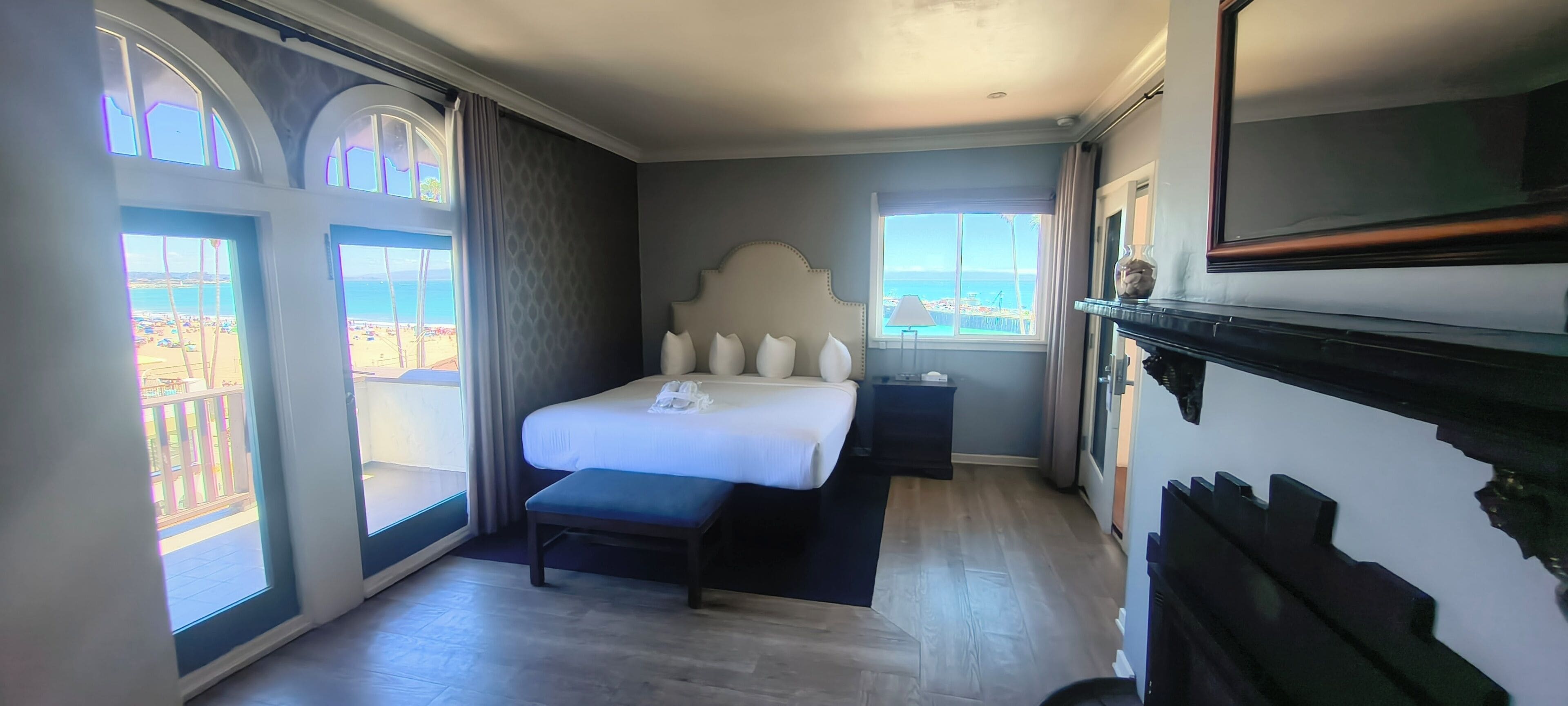 A hotel room with a bed, a bench, a TV, and large windows with a view of the beach.