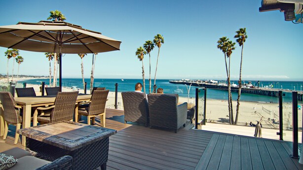 Terraza exterior con mobiliario de mimbre y una sombrilla grande, con vista a una playa y un muelle.