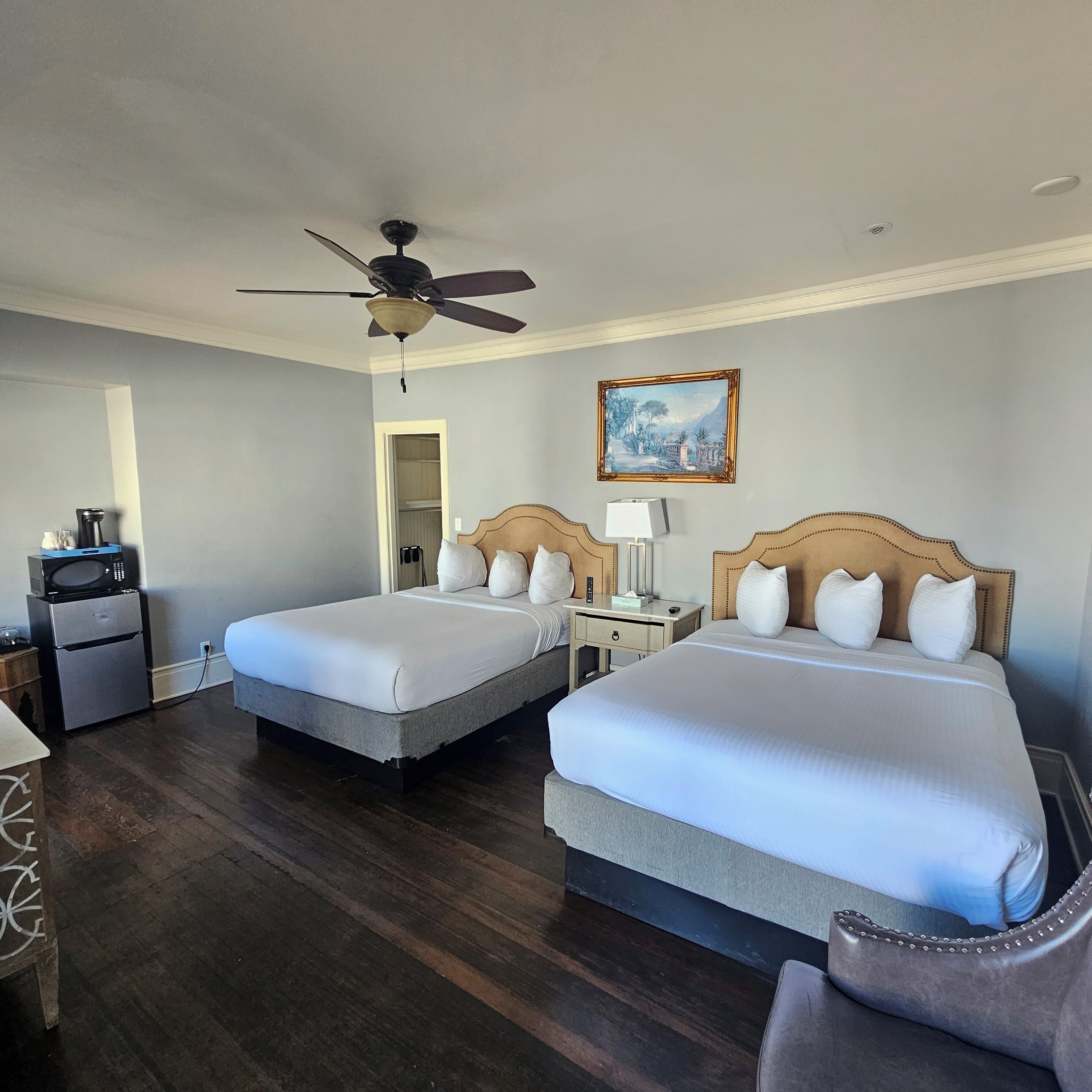 A hotel room with two beds, a ceiling fan, a painting, a microwave, and a refrigerator.