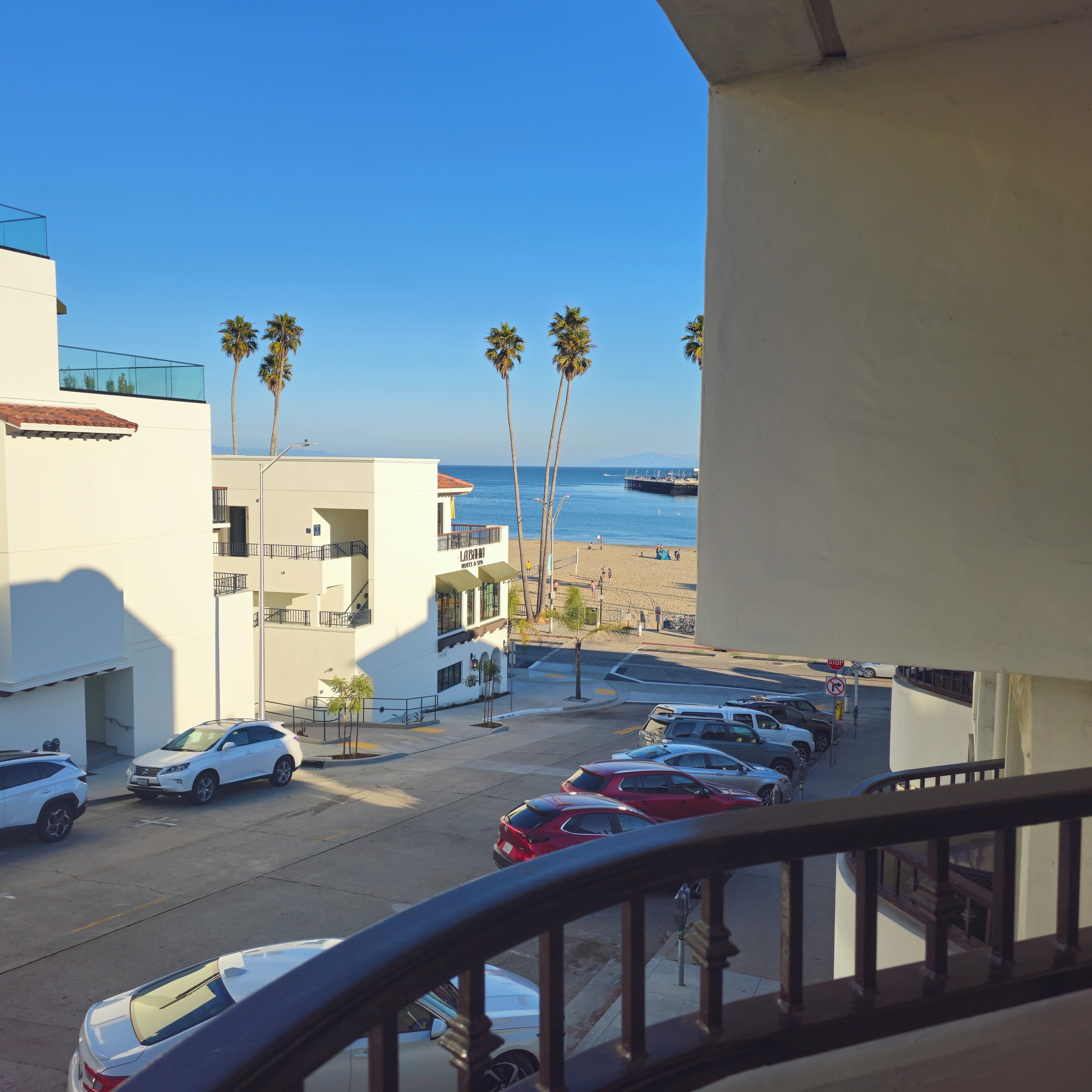 Vista da un balcone su un parcheggio con diverse auto, una spiaggia e palme all'orizzonte.