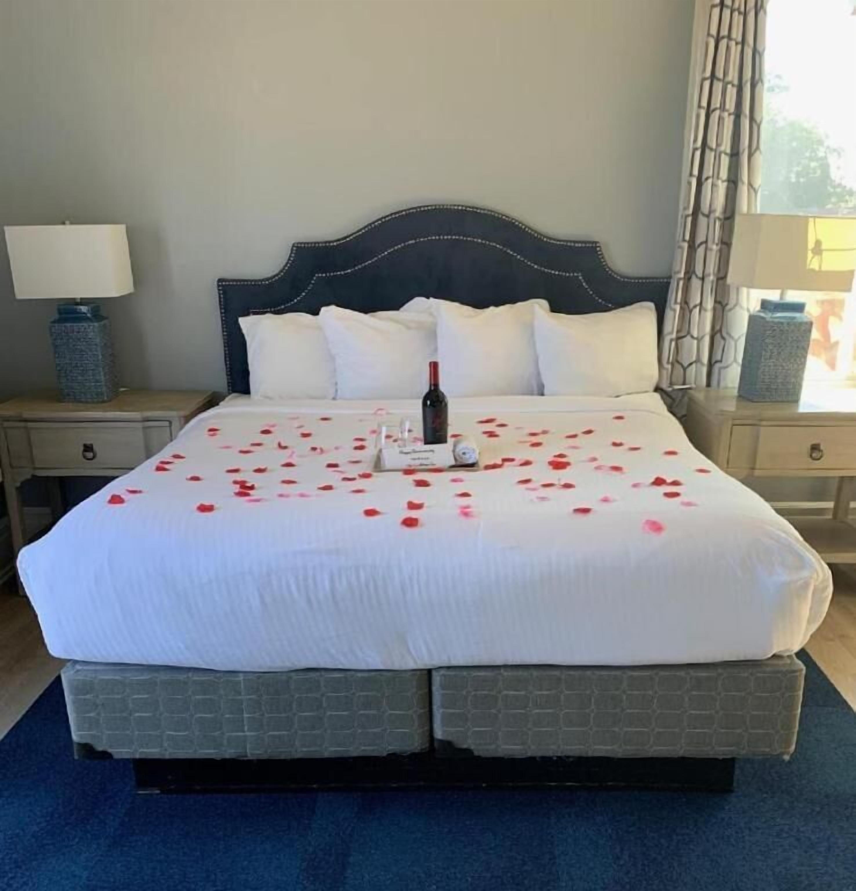 A neatly made bed with rose petals and a bottle, a dark headboard, and a lamp on a bedside table.