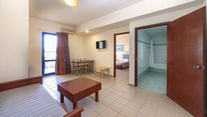 A hotel room with a bed, a wooden table, a TV, a shower area, and a balcony with curtains. - Olympic Suites Hotel Apartments (Rethymno)
