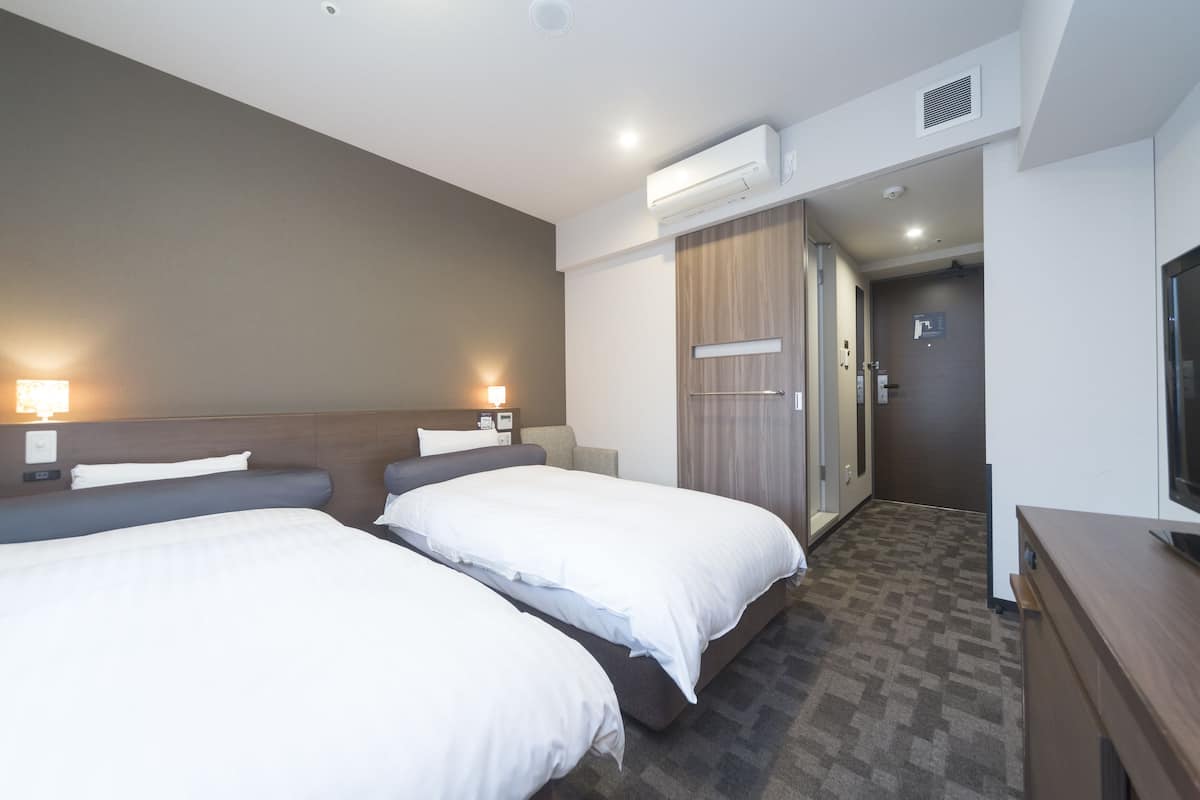 A hotel room with two beds, a TV, an air conditioning unit, and a hallway leading to a bathroom.