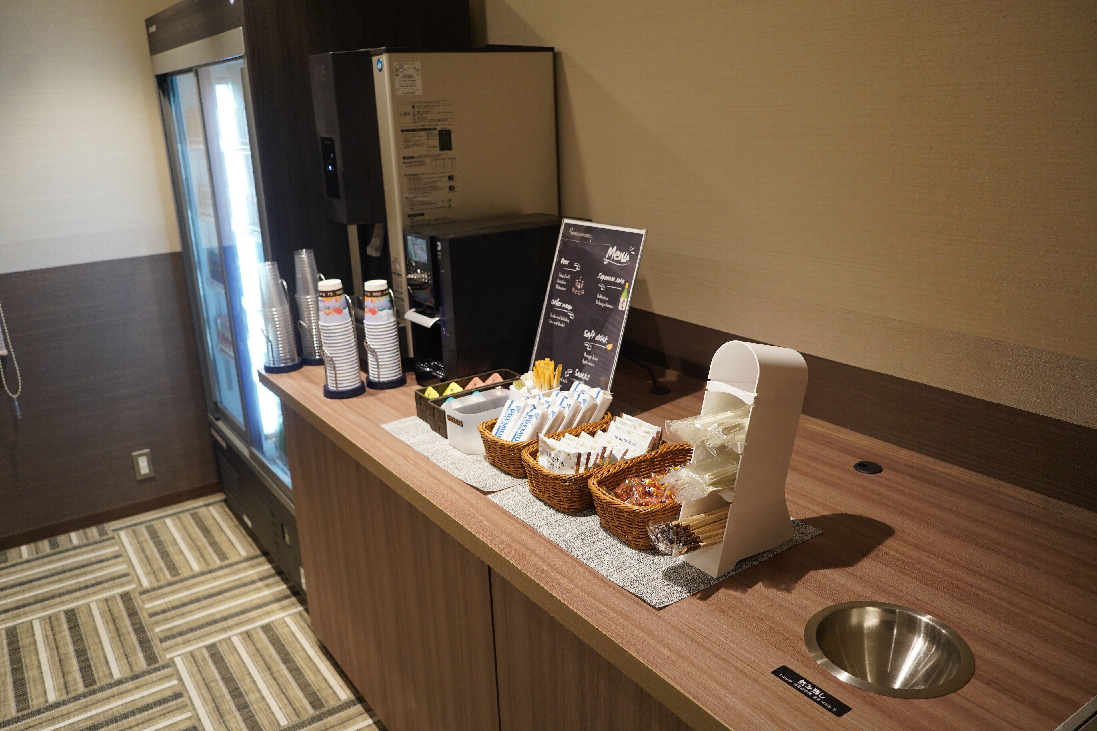 A hotel reception area with a coffee machine, a refrigerator, and a buffet counter with various food items and condiments.