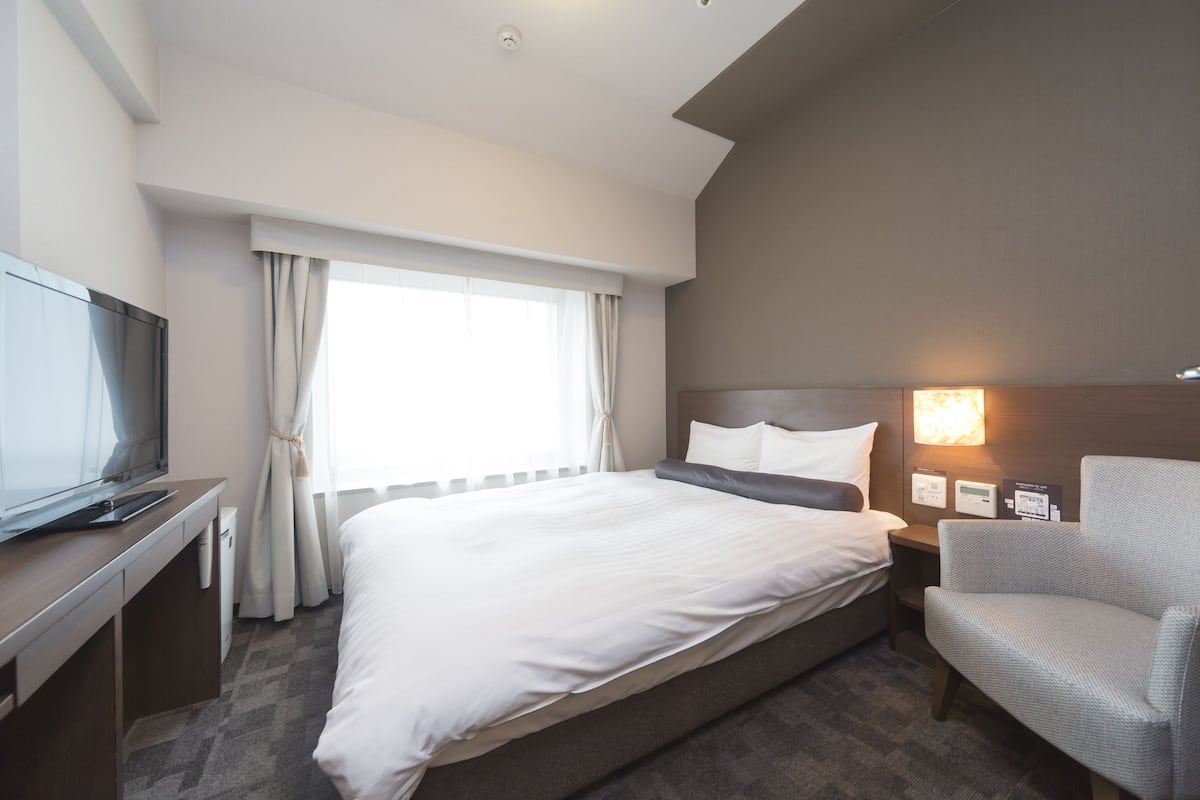 A hotel room with a large bed, a desk with a television, a chair, and a nightstand with a lamp.