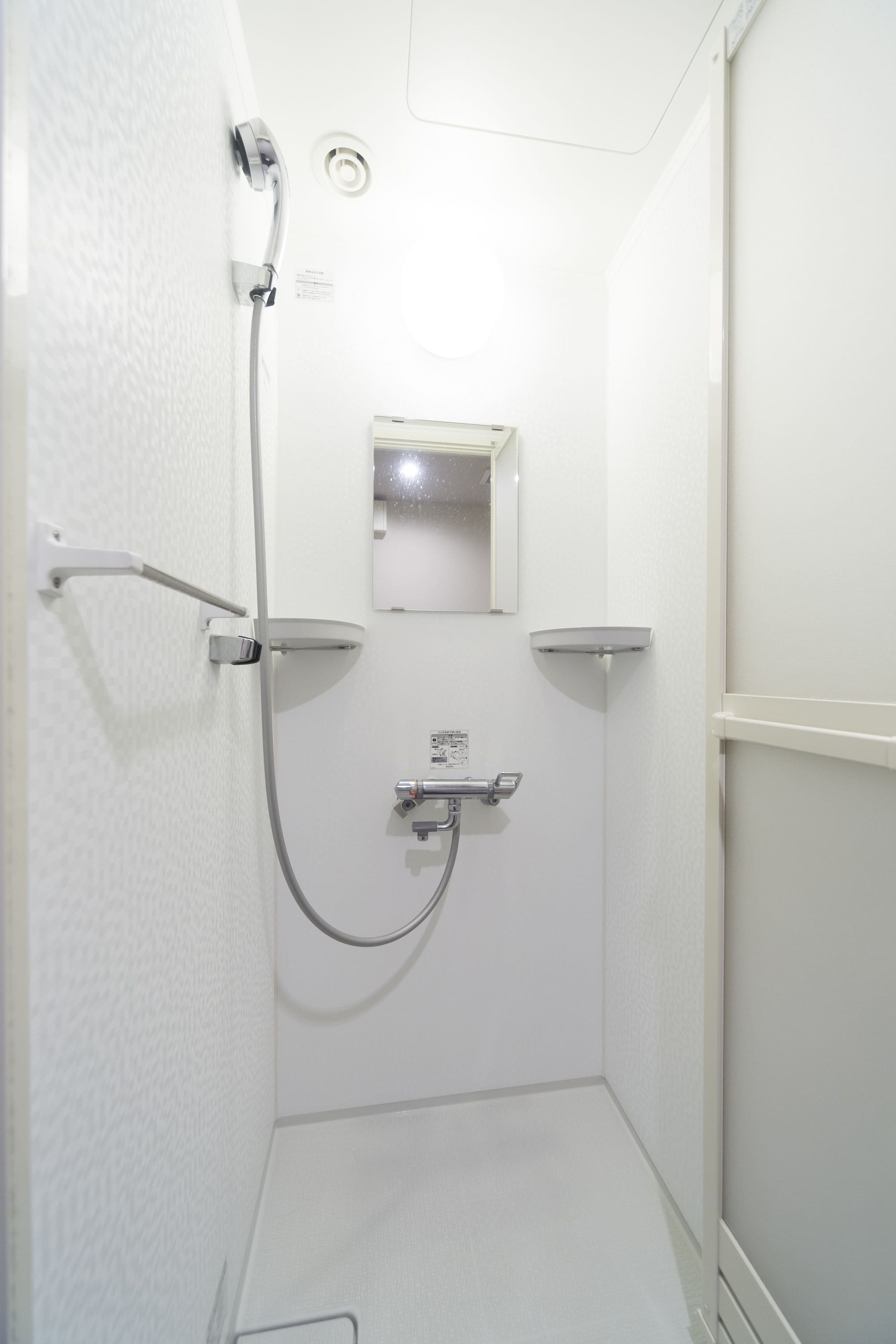 Un petit espace de douche fermé, de couleur blanche, équipé d’un pommeau de douche à main, d’un miroir fixé au mur et d’un panneau de commande.