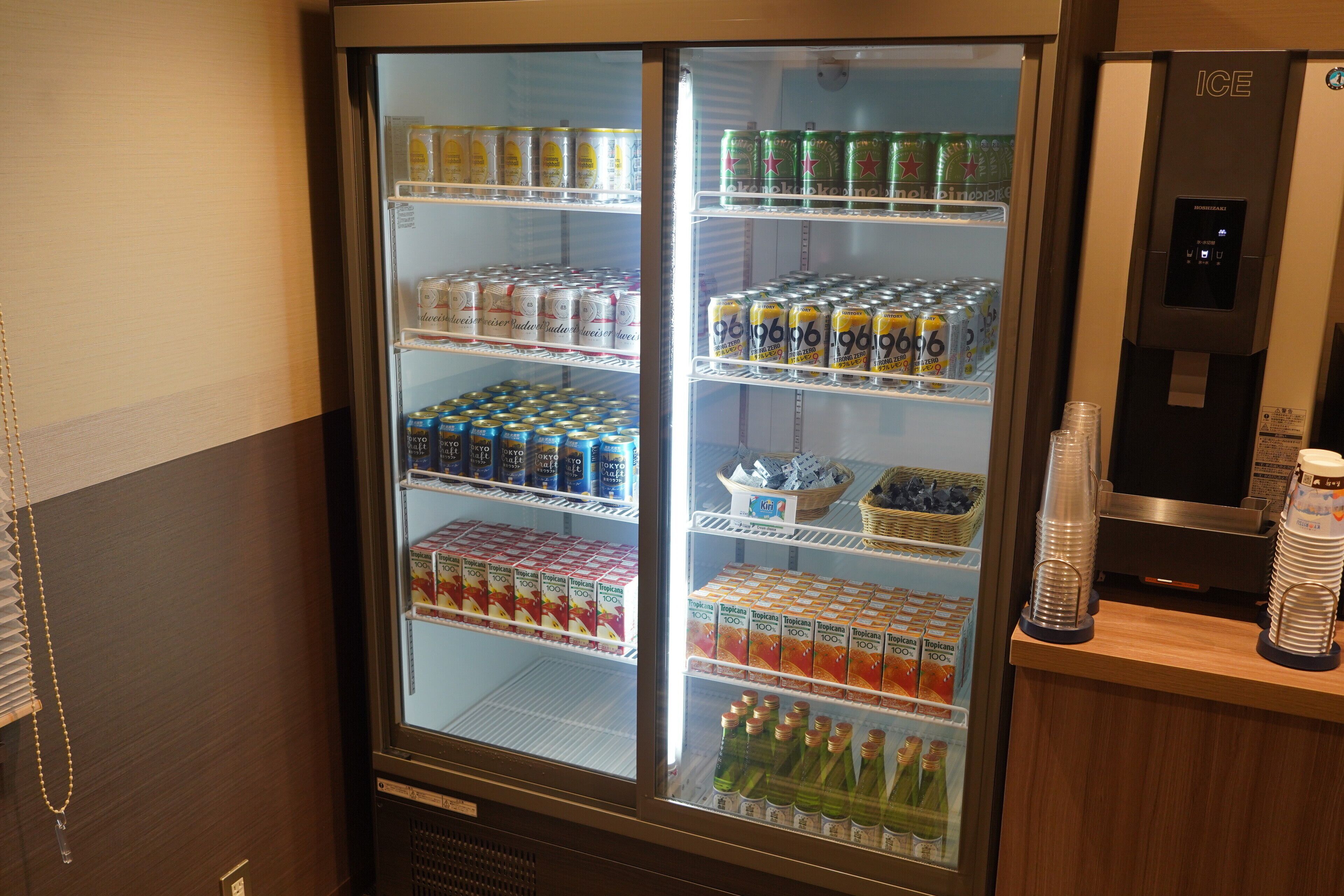 Un refrigerador de bebidas repleto de diversas opciones, incluyendo latas y botellas, y un dispensador de agua a la derecha.