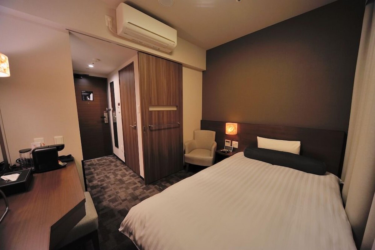 A hotel room with a bed, a desk, a chair, an air conditioning unit, and a door leading to a bathroom.