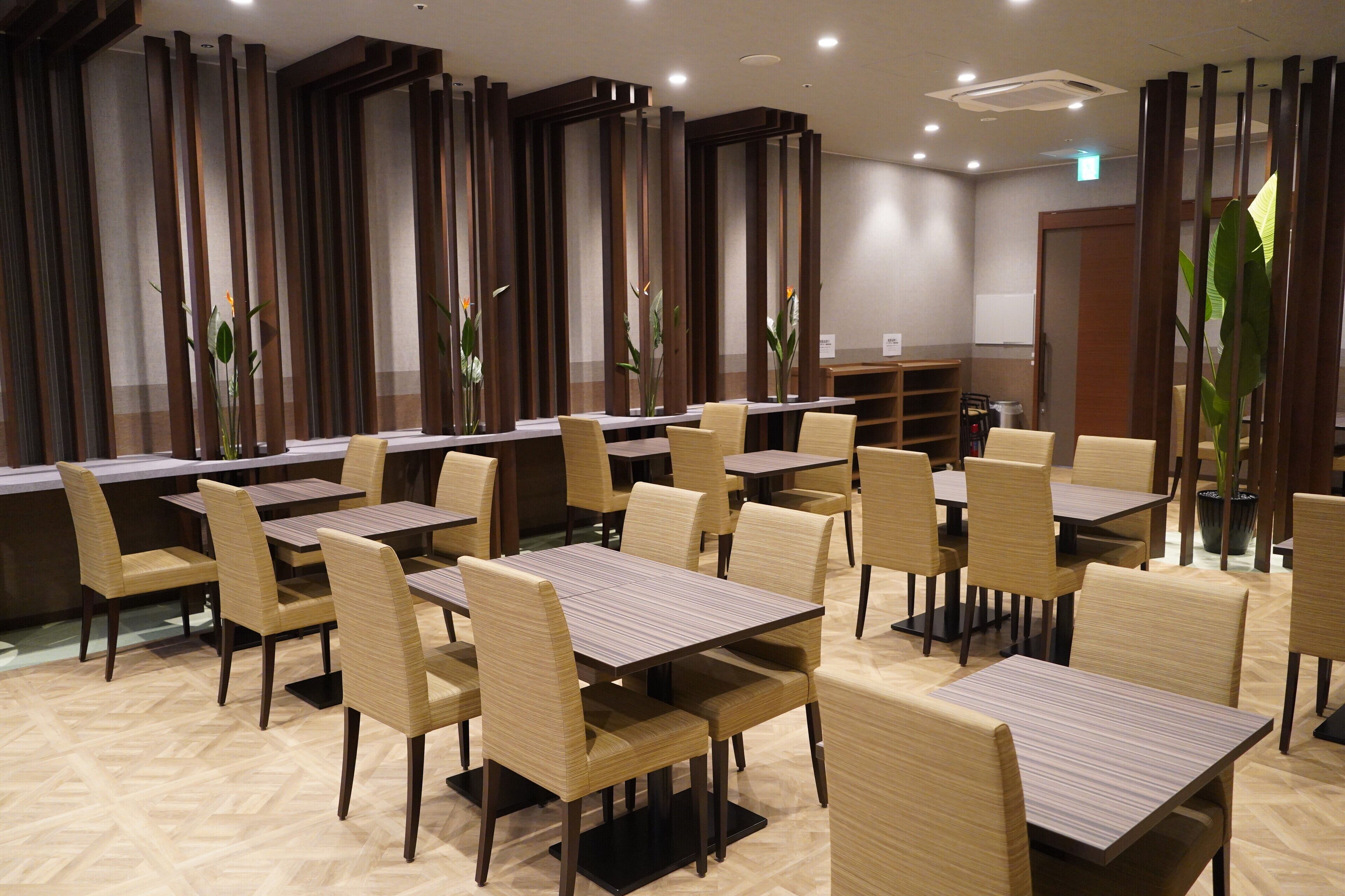 A hotel dining area with multiple tables and chairs, wooden partitions, and potted plants.