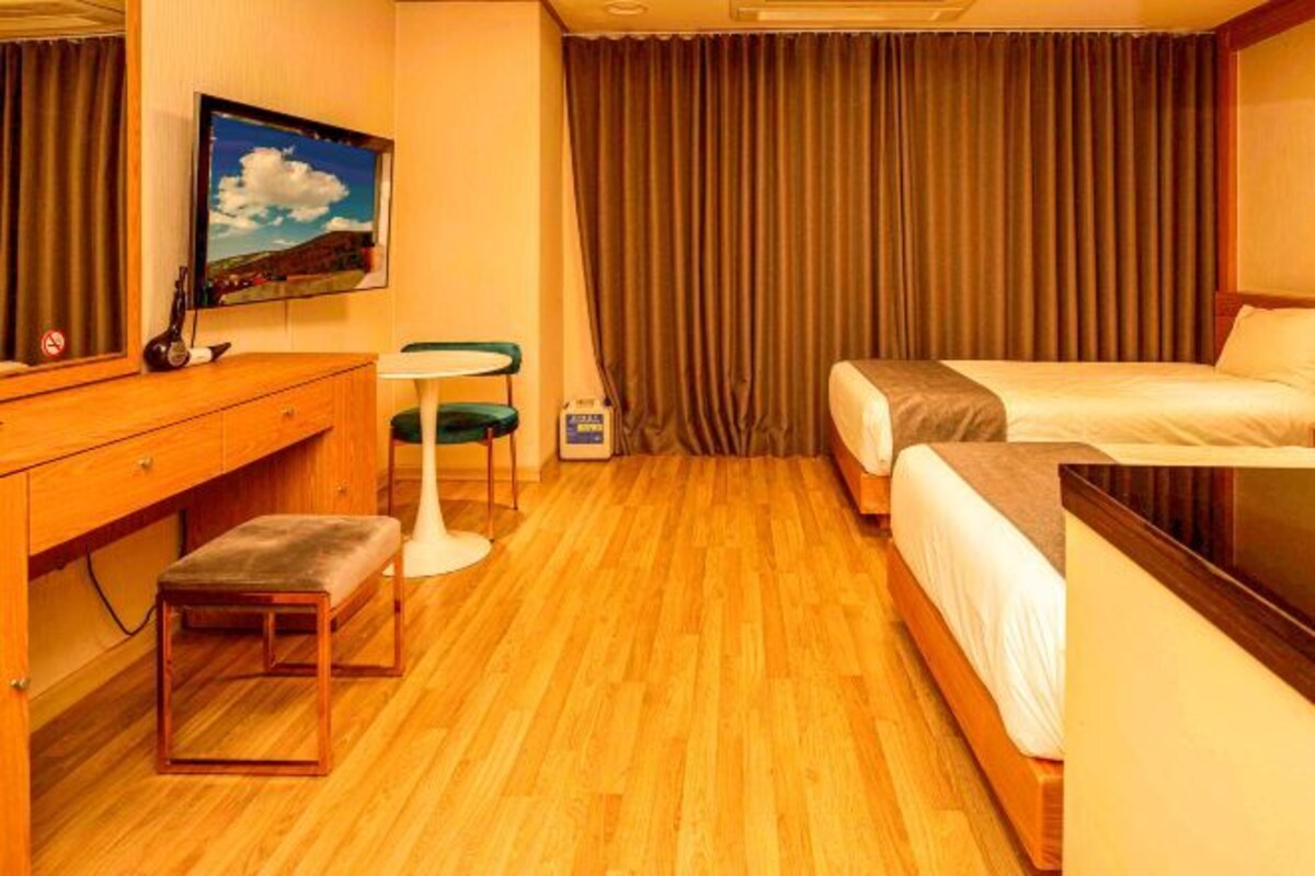 Una habitación de hotel con suelo de madera, un escritorio con una silla, un banco, una cama y un televisor montado en la pared.
