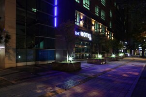 A hotel with a modern facade, illuminated signage, and landscaped planters along the walkway.