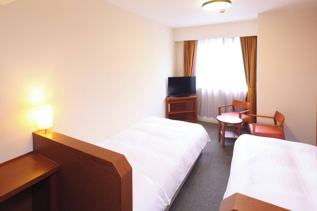 A hotel room with two single beds, a desk with a lamp, a television on a stand, and a chair.
