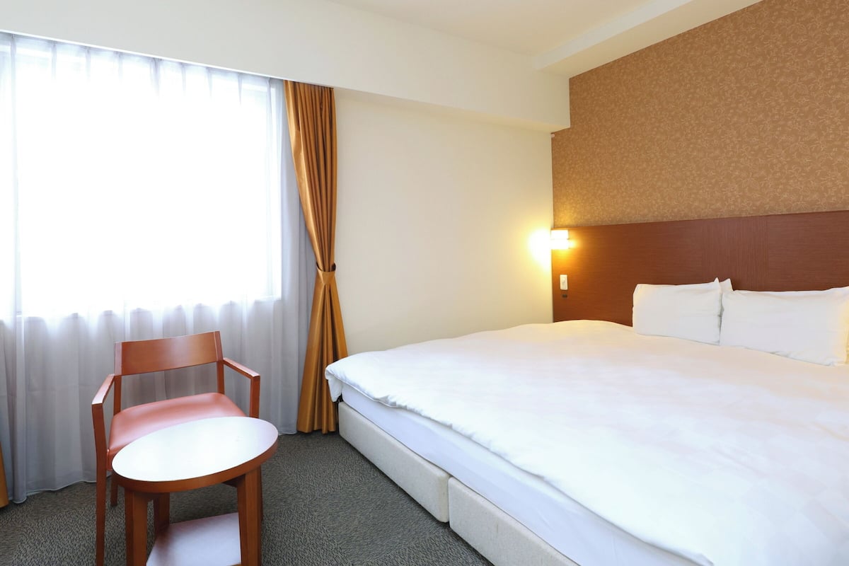 A hotel room with a bed, a chair, a nightstand, and a window with curtains.