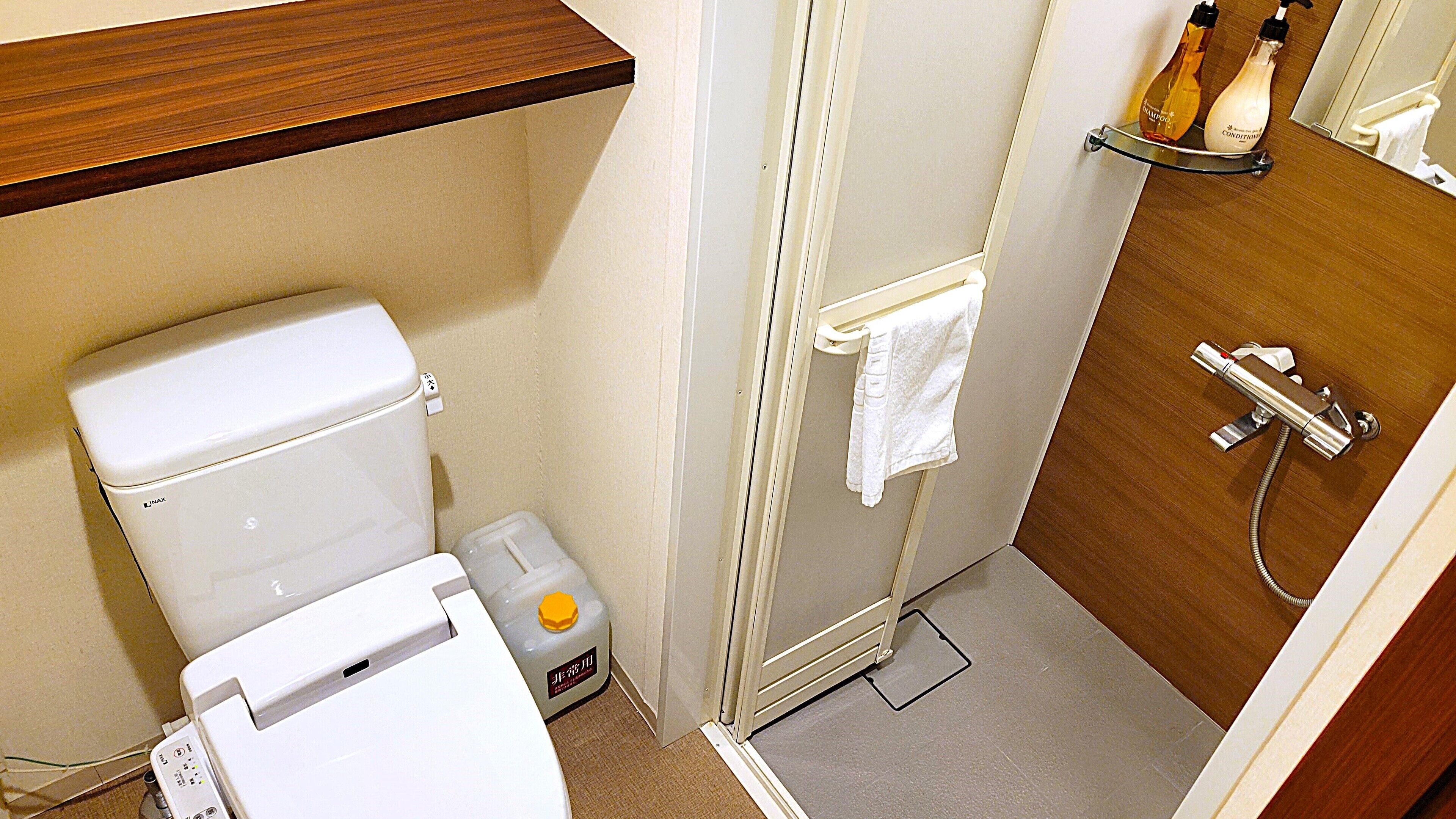 A compact bathroom with a toilet, a small trash bin, a shower area, and a shelf with toiletries.