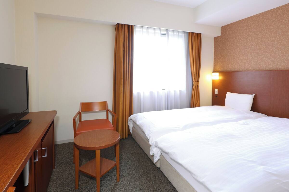 A hotel room with a bed, a desk with a television, a chair, and a window with curtains.