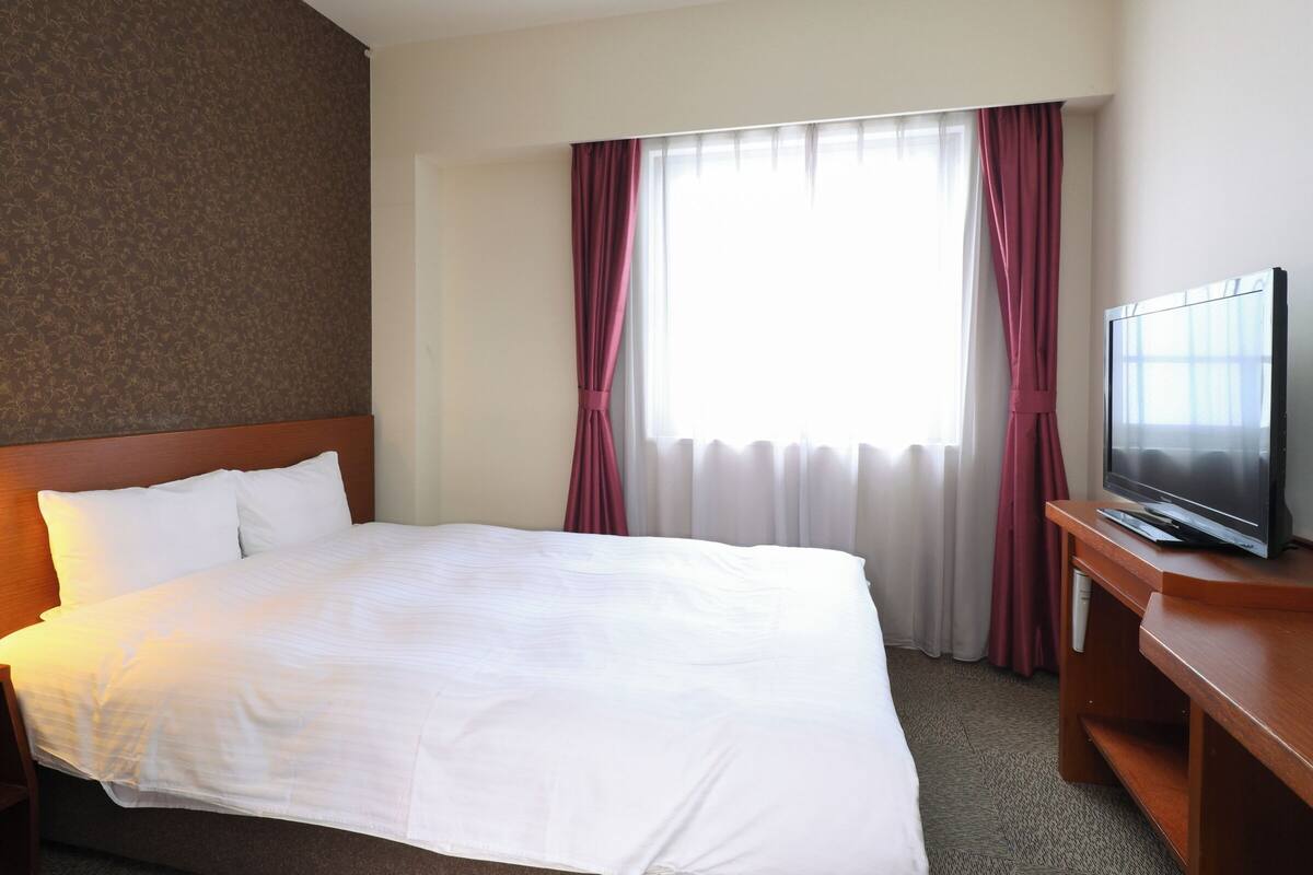 A hotel room with a large bed, a television on a wooden stand, and a window with curtains.