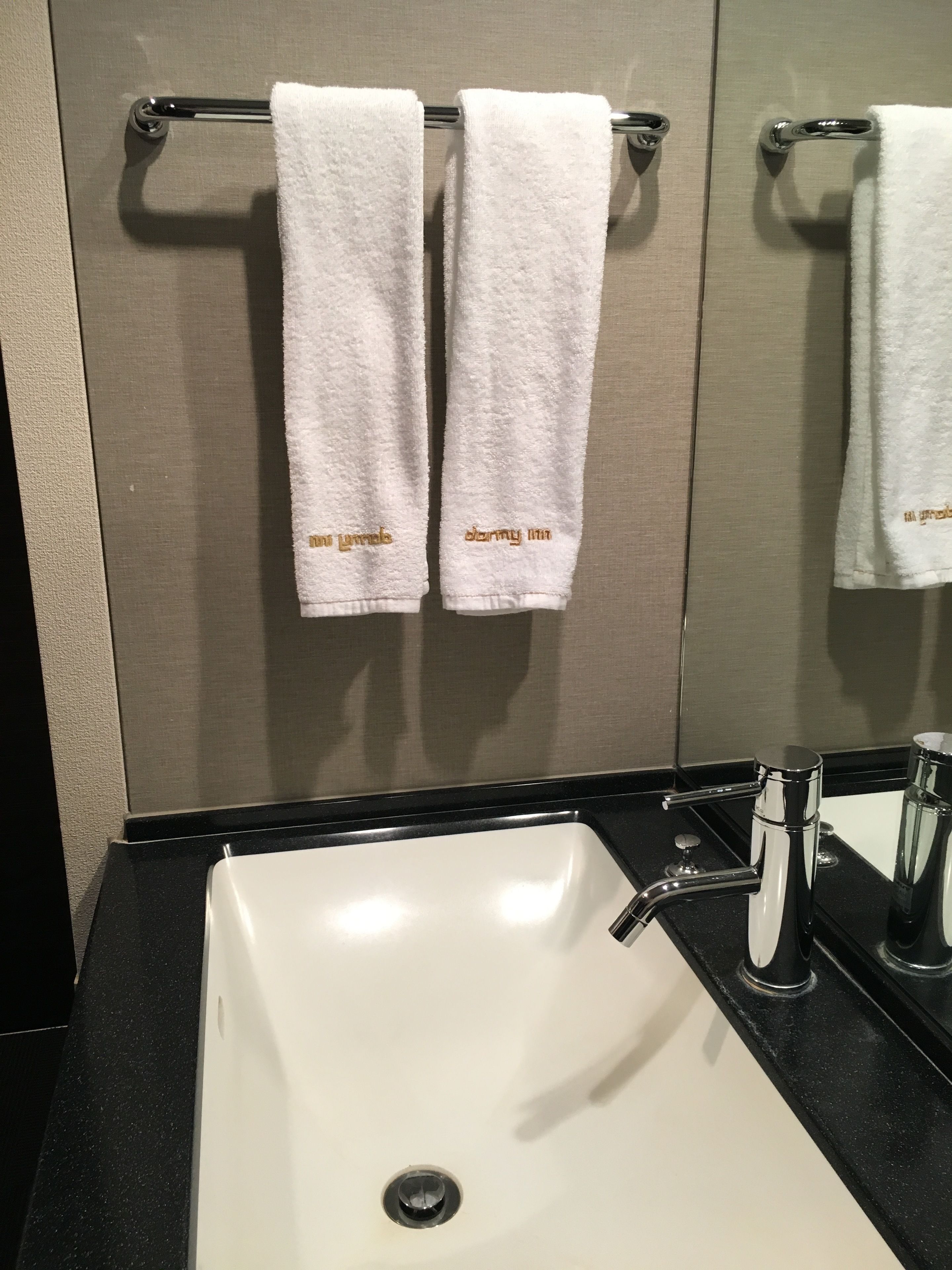 Un baño con un lavamanos blanco, un espejo y dos toallas blancas colgadas en un perchero.