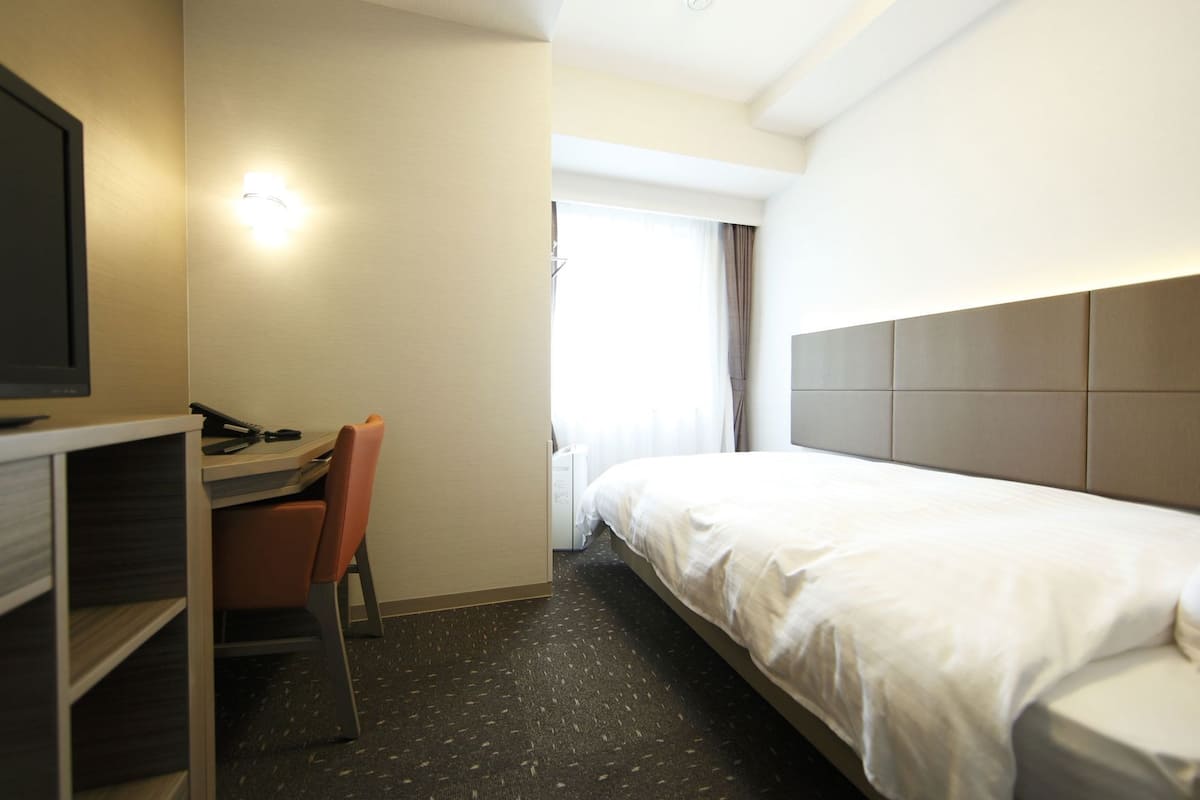 A hotel room with a bed, a desk with a chair, a television, and a window with curtains.