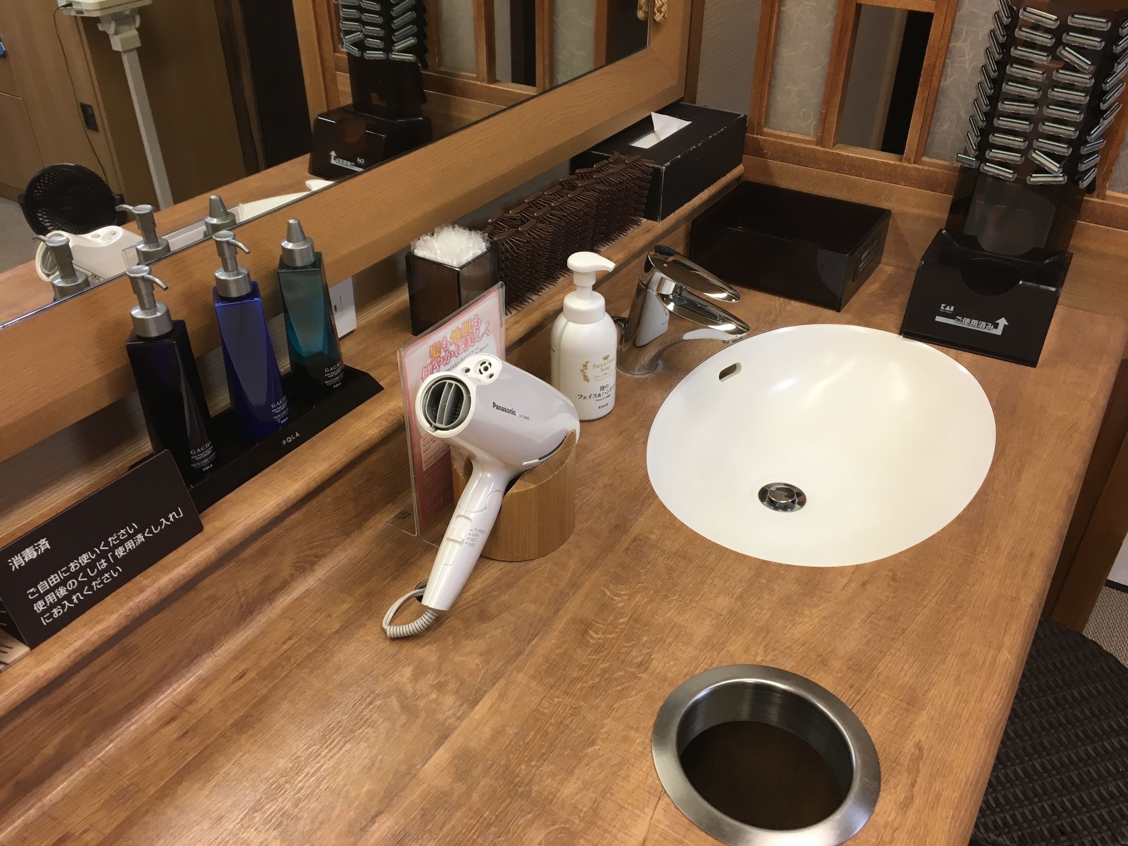 A bathroom with a wooden vanity, a white sink, a Panasonic hair dryer, and various toiletries.