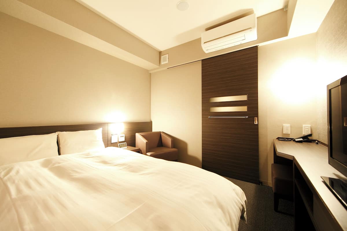 A hotel room with a large bed, a desk, a chair, a TV, and an air conditioning unit.