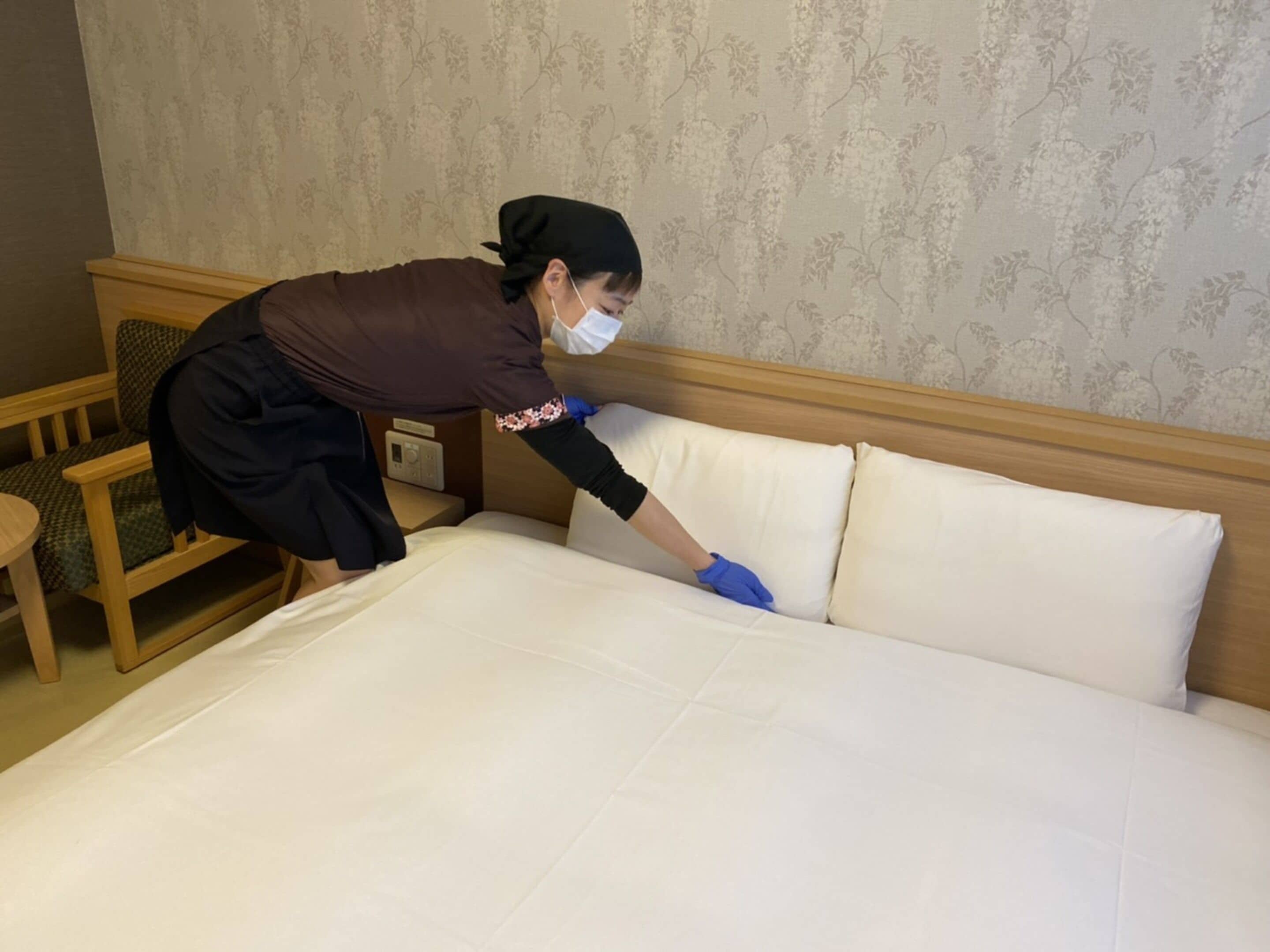 A person cleaning a bed in a hotel room with floral wallpaper and wooden furniture.