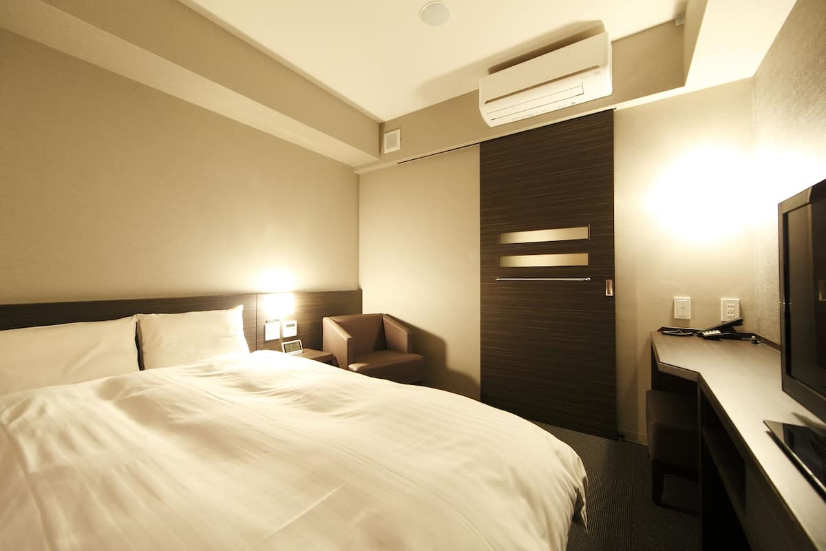 A hotel room with a large bed, a desk, a chair, a TV, and an air conditioning unit.