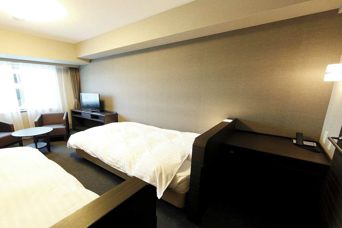 A hotel room with two beds, a desk, a chair, a TV, and a window with curtains.