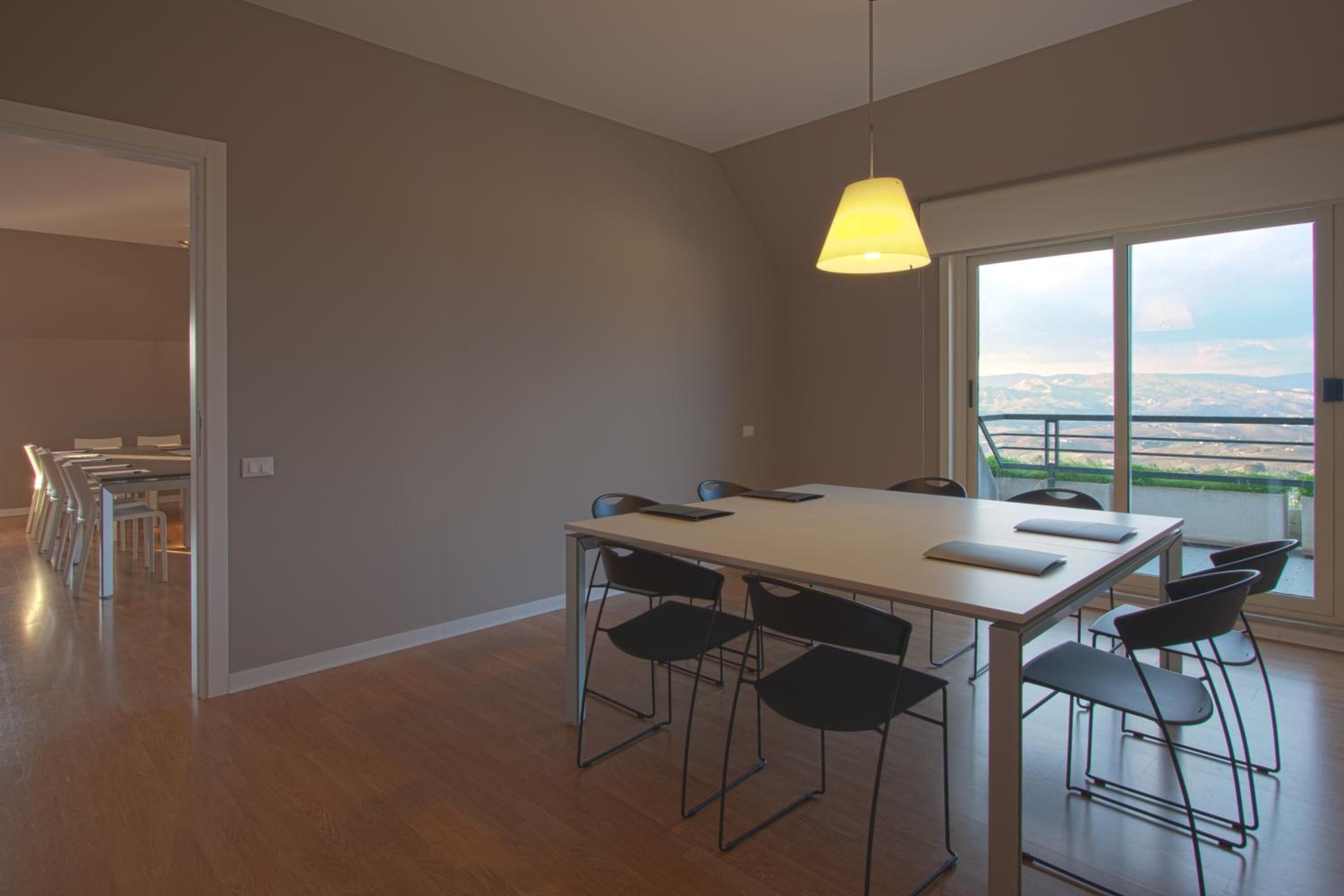 A modern dining area with a large table and chairs, a hanging light fixture, and a view through large windows.