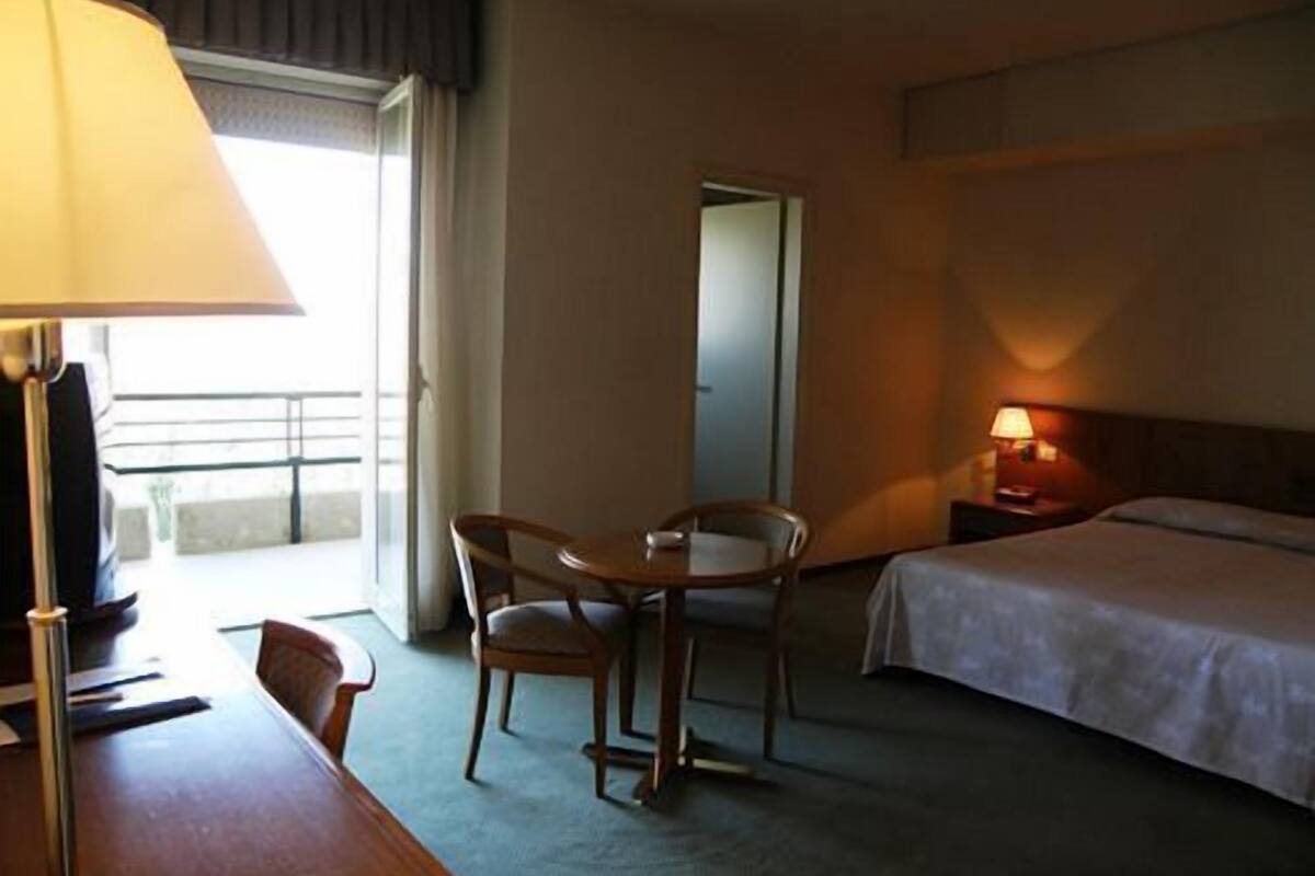 A hotel room with a bed, a desk with a lamp, a chair, and a view of the outside balcony.