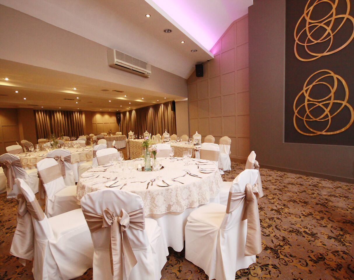 Une salle de banquet avec des tables rondes dressées pour un événement formel, des chaises blanches ornées de nœuds et un mur décoratif aux motifs abstraits.
