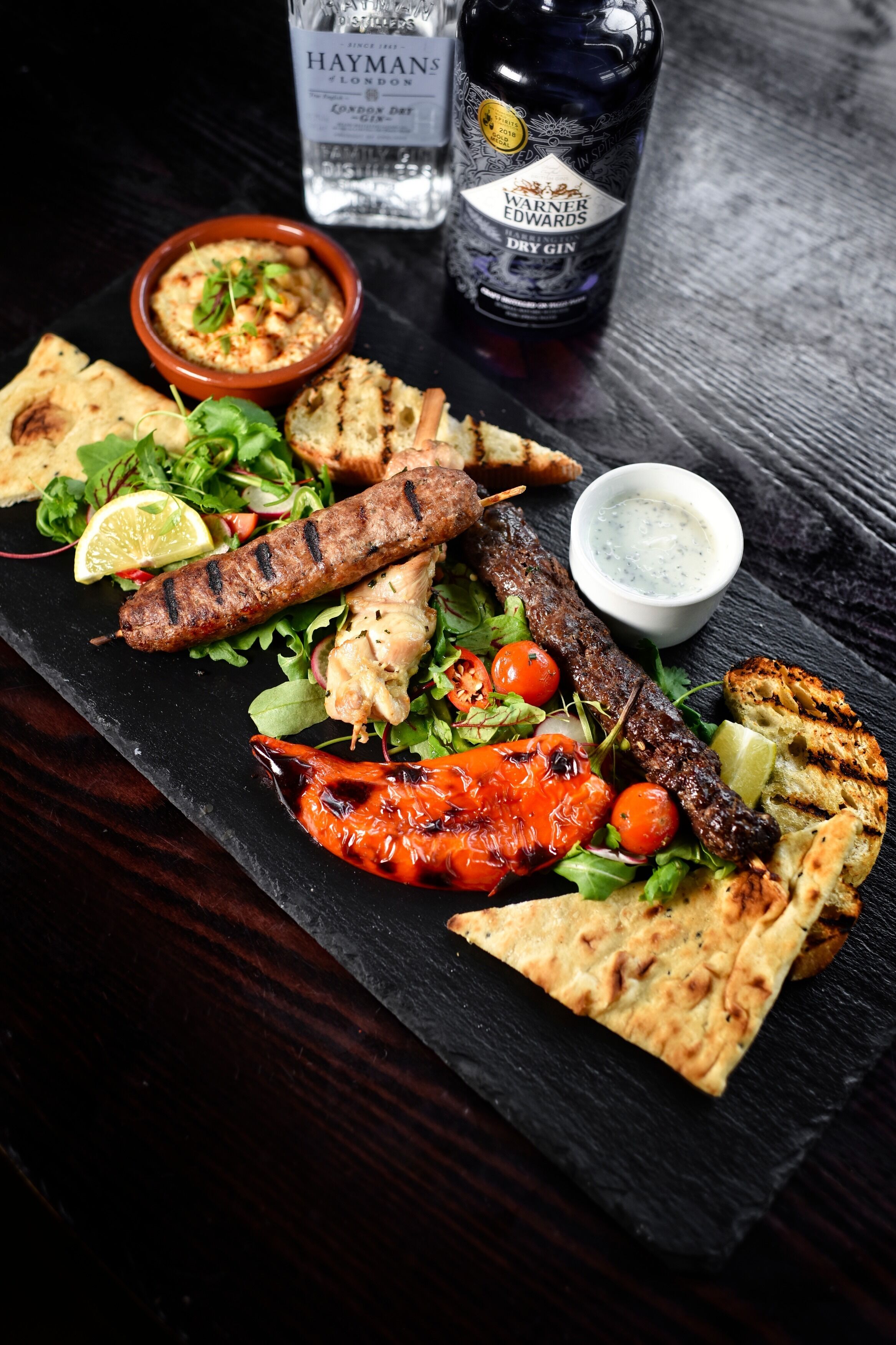 A variety of grilled meats, vegetables, and flatbread on a slate board, accompanied by a bottle of dry gin and a small bowl of sauce.