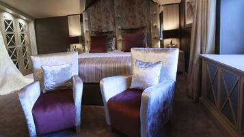 A bedroom with a large bed, two upholstered chairs, a wardrobe, and a window.