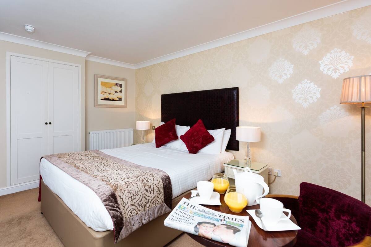 Quarto de hotel com cama, mesas de cabeceira, uma cadeira, abajur e bandeja de café da manhã com um jornal.