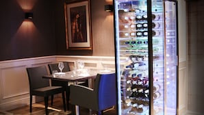 Un área de comedor con una mesa lista para dos personas, un enfriador de vinos con botellas y un cuadro en la pared.