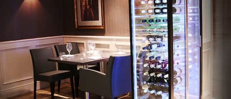A dining area with a table set for two, a wine cooler with bottles, and a framed picture on the wall.