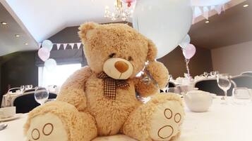 A plush teddy bear on a dining table with balloons and a chandelier in the background.