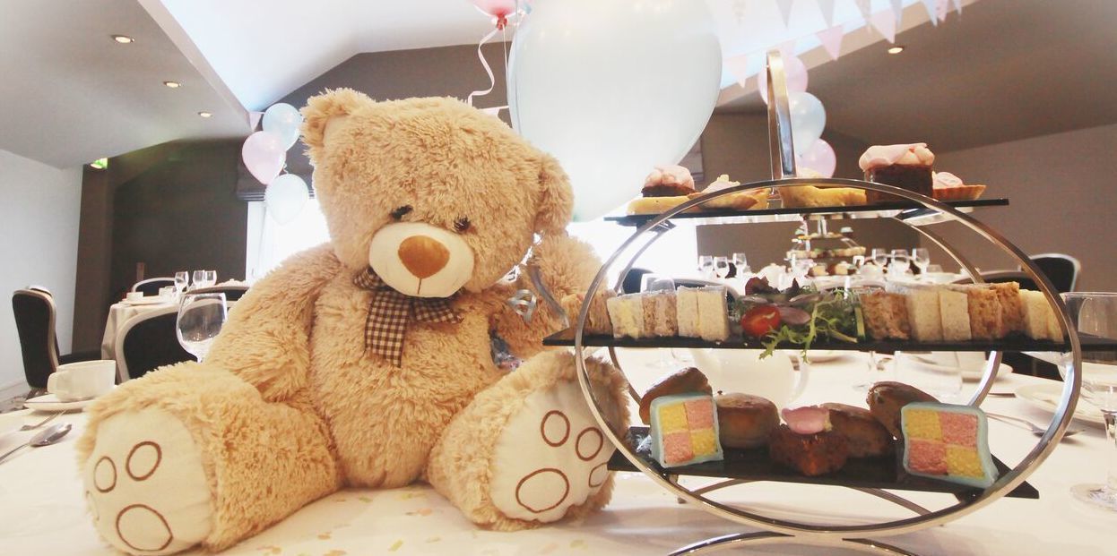A teddy bear seated next to a dessert display at a dining area with balloons in the background.