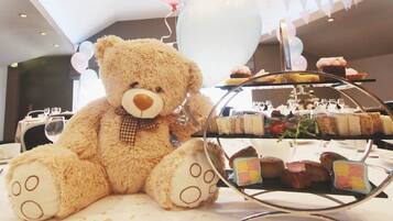Un osito de peluche sentado junto a una mesa de postres en un área de comedor, con globos al fondo.