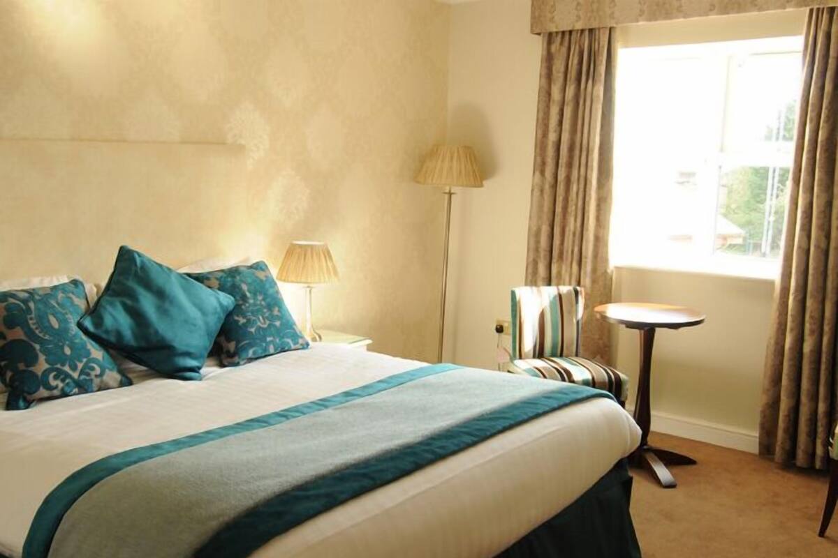 Quarto de hotel com cama, luminária, cadeira, mesa pequena e janela com cortinas.
