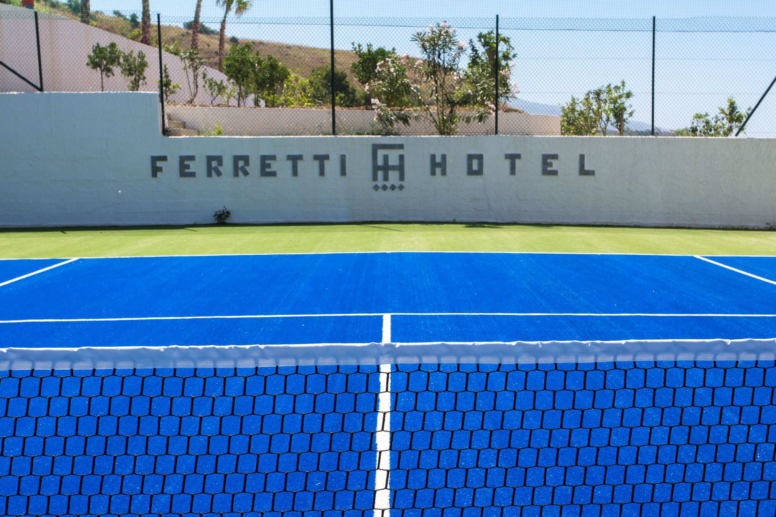 A tennis court with a net in the foreground and a wall with the text "FERRETTI HOTEL" in the background.