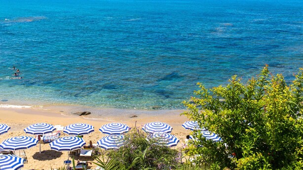 Une plage avec une eau d’un bleu limpide, plusieurs parasols rayés de bleu et de blanc, et quelques chaises de plage.
