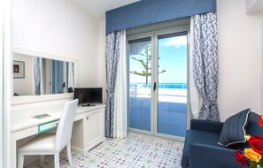A hotel room with a white desk, a chair, a television, a sofa with a pillow, and a view of the sea through large glass doors. - Hotel Ferretti (Diamante)