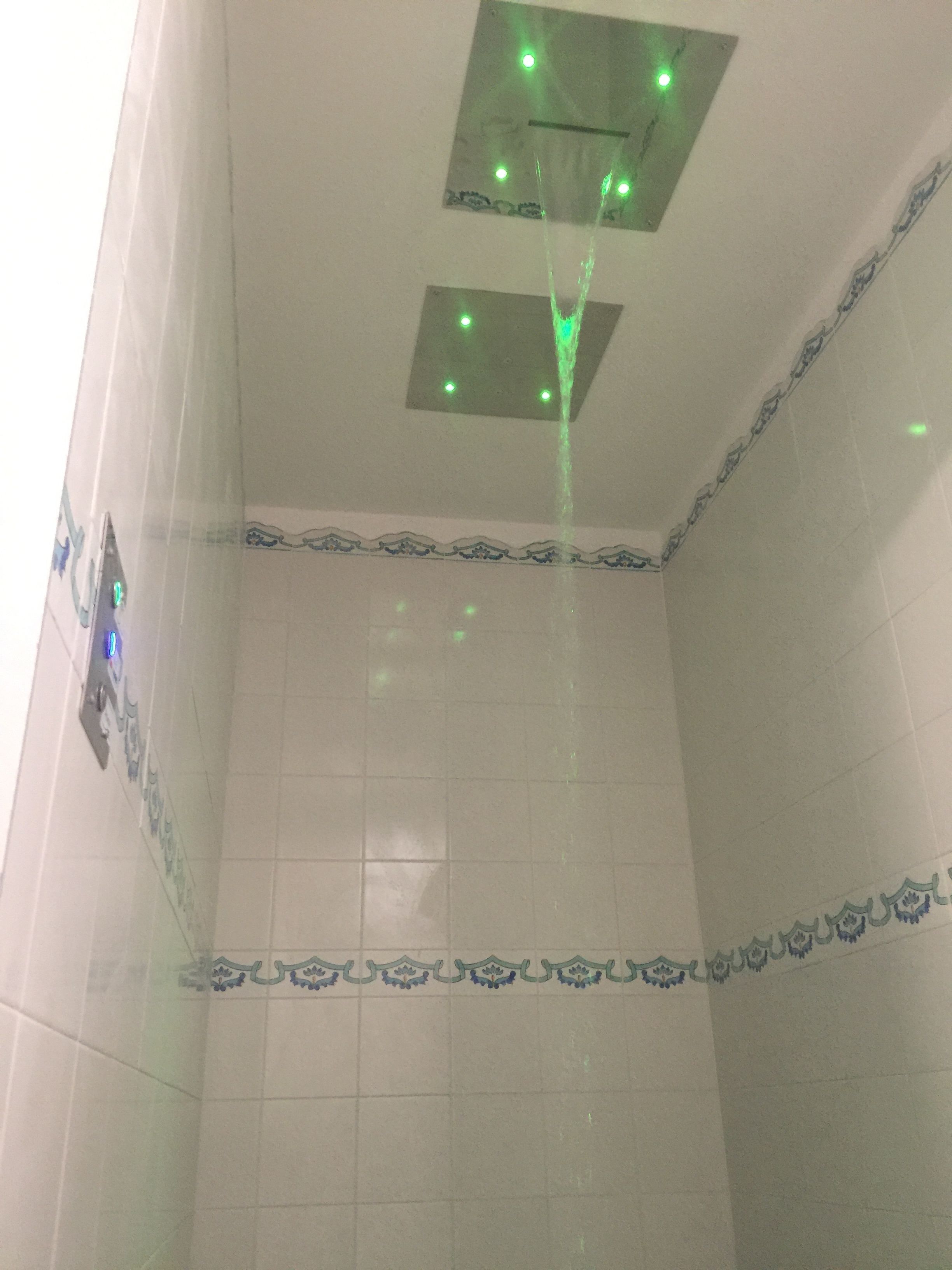 Baño con azulejos blancos y un borde decorativo azul. Hay dos lámparas cuadradas en el techo, una de las cuales emite luz verde.