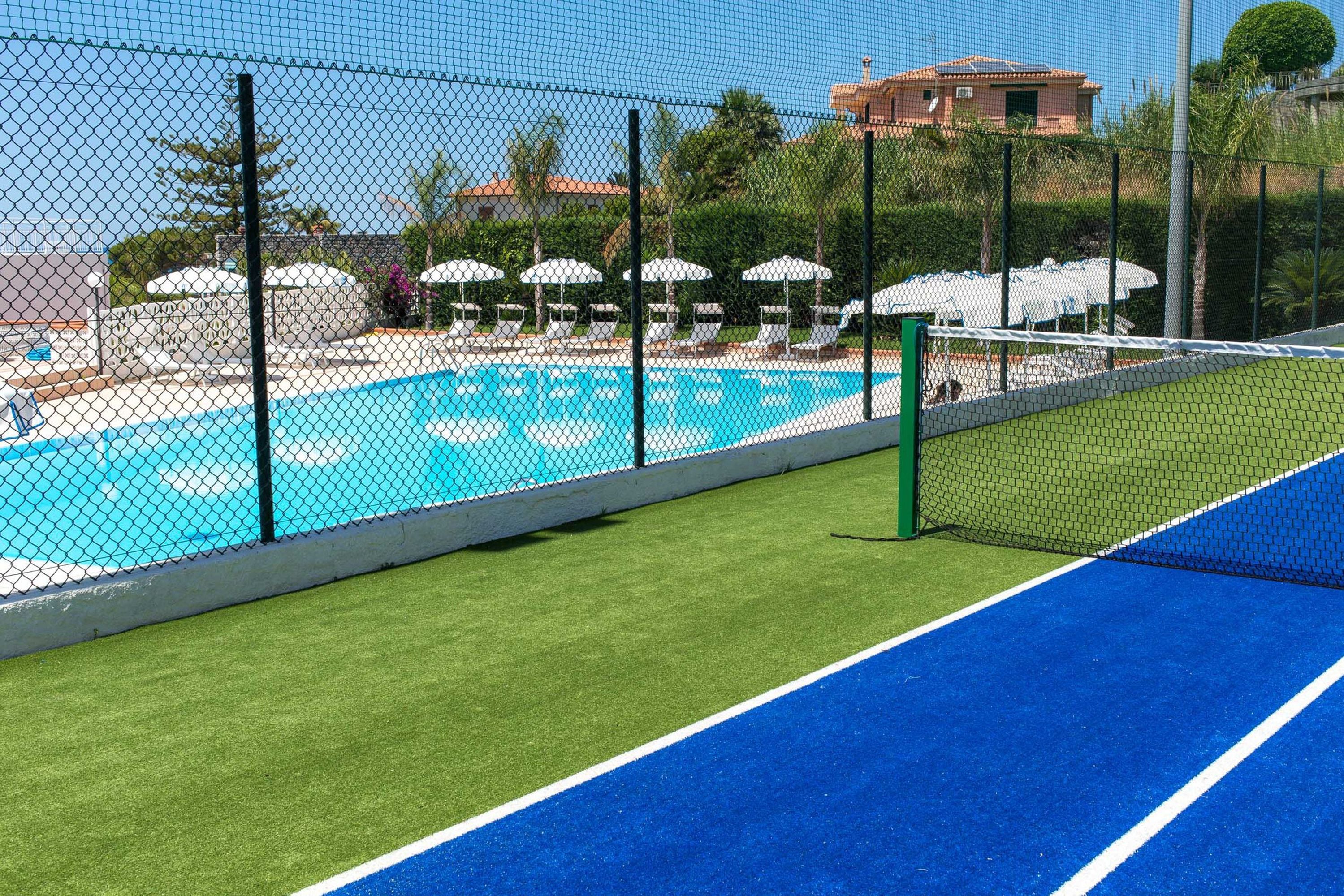 A tennis court adjacent to a swimming pool with lounge chairs and umbrellas.