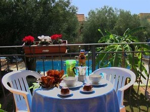 A balcony with a table set for tea, surrounded by chairs, with a view of a swimming pool and greenery.