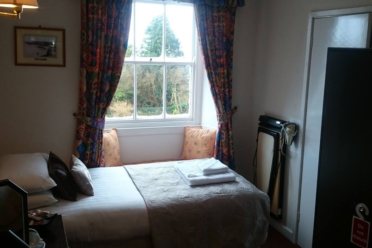Una habitación de hotel con una cama, una ventana con cortinas, una lámpara de noche, un radiador y una puerta con un cartel de 'No molestar'.